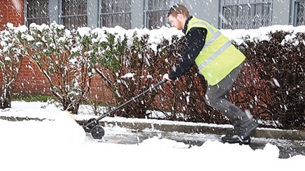 A man wearing personal protective equipment which includes a reflective yellow hi-vis vest and black safety boots. He is shoveling a large amount of snow outside using a yellow and black One-Man Snow Plough.