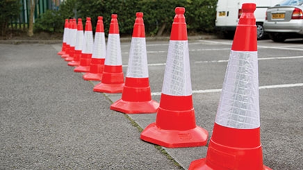 Ten red JSP® Sand Weighted One Piece Traffic Cones with high visibility sleeves are in a car park in a row horizontally restricting access to certain parking bays.