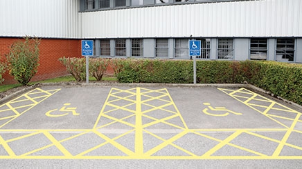 A car park which has two designated disabled parking bays. The disabled parking bays have been created using yellow line marking paint and there are two blue and white ‘disabled parking only’ car park signs on a grey post in front of the parking bays.