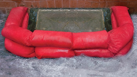 Two red HydroSnake Flood Prevention barriers are being used to stop flood water entering a drain.
