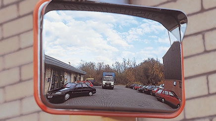 A red Anti-Vandal Traffic Mirror has been post mounted in a car park. In the reflection of the mirror you can see many cars parked and a large lorry parked in the middle of the car park.